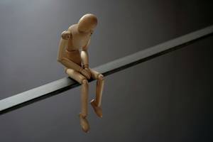 Holzfigur sitzt mit gesenktem Kopf und verschränkten Händen auf einer Kante