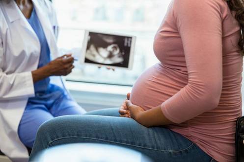Schwangere Frau sitzt auf einer Untersuchungsliege, im Hintergrund zeigt eine Ärztin ein Ultraschallbild